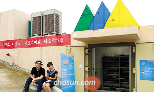 백령도 제7호 대피소 외벽에 '나오쇼, 나오쇼, 나오지 마쇼, 나오지 마쇼'라 적힌 플래카드는 미술가 이지수의 작품 '공격2'. 현관 지붕에 설치된 녹색·파랑·노랑의 삼각형은 긴박함을 부각시키기 위한 김주은의 작품 'Here1'이다